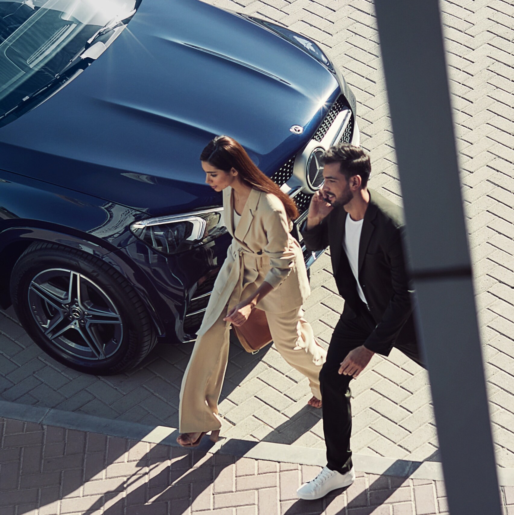 Eine Frau und ein Mann gehen an ihrem Mercedes-Benz vorbei und sind mit dem Smartphone per Digitalem Extra mit dem Fahrzeug verbunden.